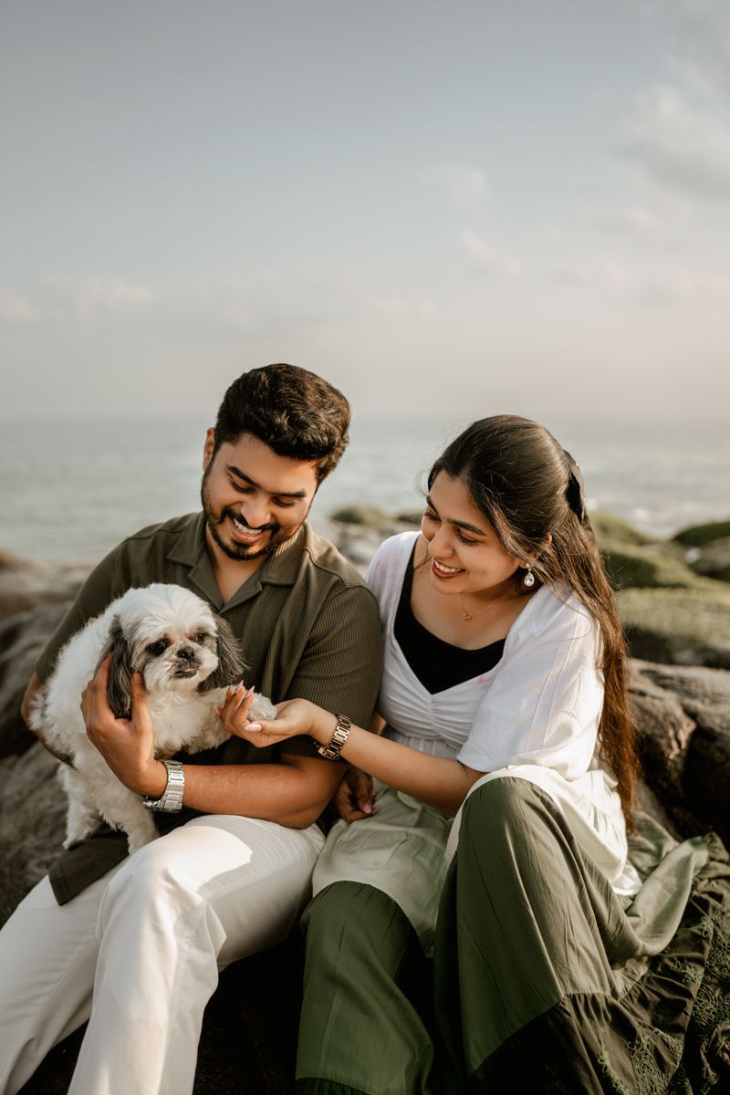A couple enjoying a tender moment with their white Shih Tzu dog on rocky coastal terrain.