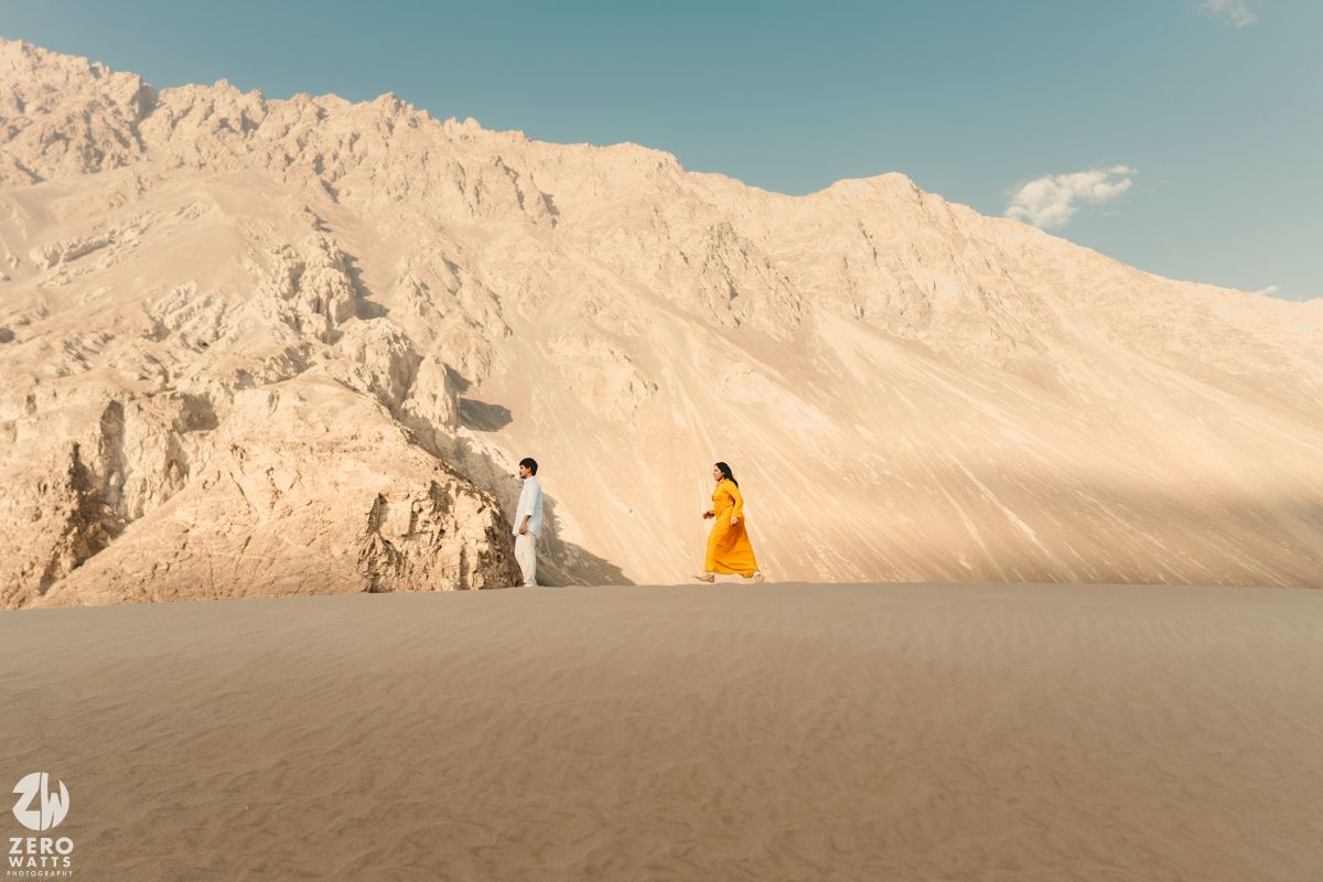 A person in a yellow dress stands on a sandy slope against towering beige mountains under a blue sky.