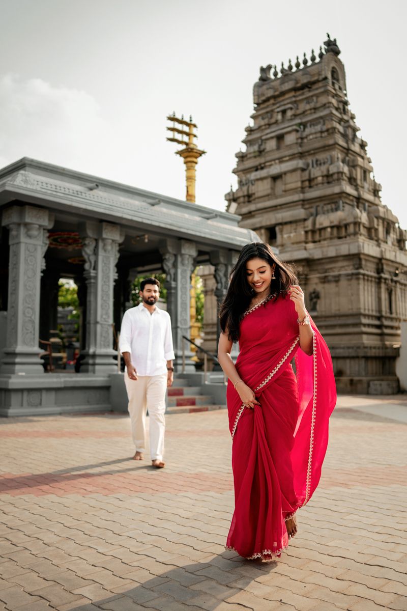 A woman in red saree twirls while a man in white stands near temple pillars and ornate architecture.