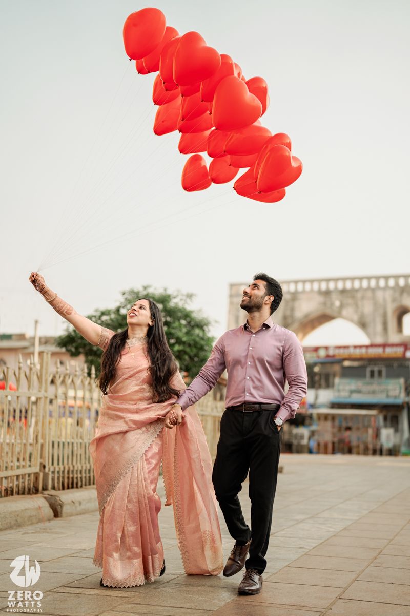 A couple in traditional attire releasing red heart-shaped balloons near a historic monument.