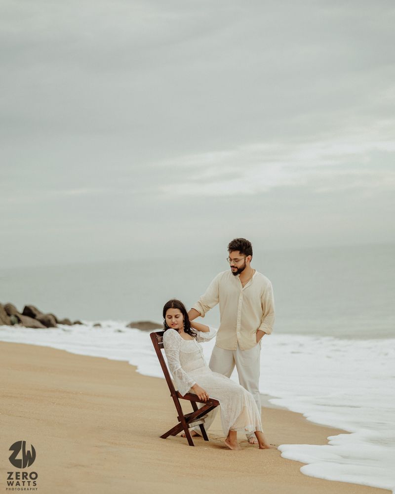 Couple in light colored outfits sitting on sandy beach during overcast day.