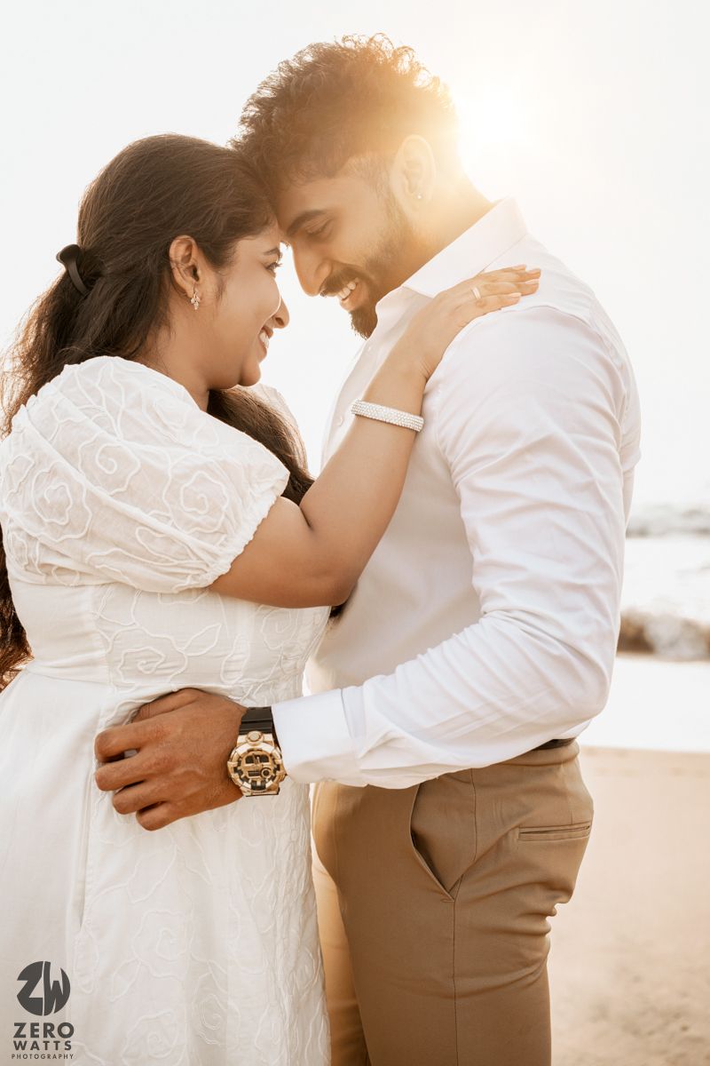 Romantic couple embraces on a bright beach in white attire during sunset photoshoot.