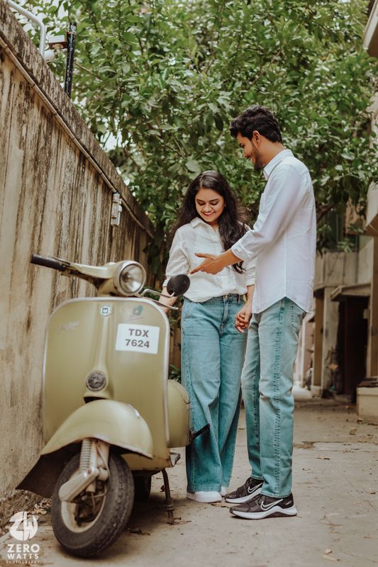 A couple standing by a vintage Vespa scooter in casual wear on a tree-lined street.