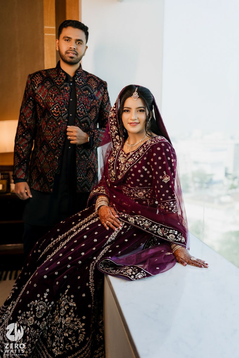 A couple in traditional South Asian wedding attire pose elegantly against a bright window backdrop.