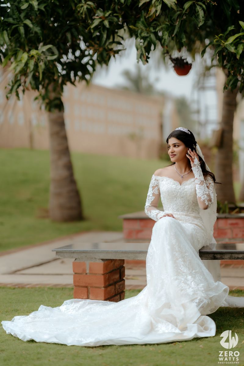 A bride in a stunning off-shoulder lace wedding gown with long sleeves poses elegantly in a garden setting with palm trees.