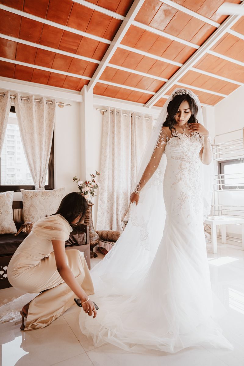 Helper assists with adjusting a long white lace wedding gown with sheer long sleeves in a bright sunlit room.