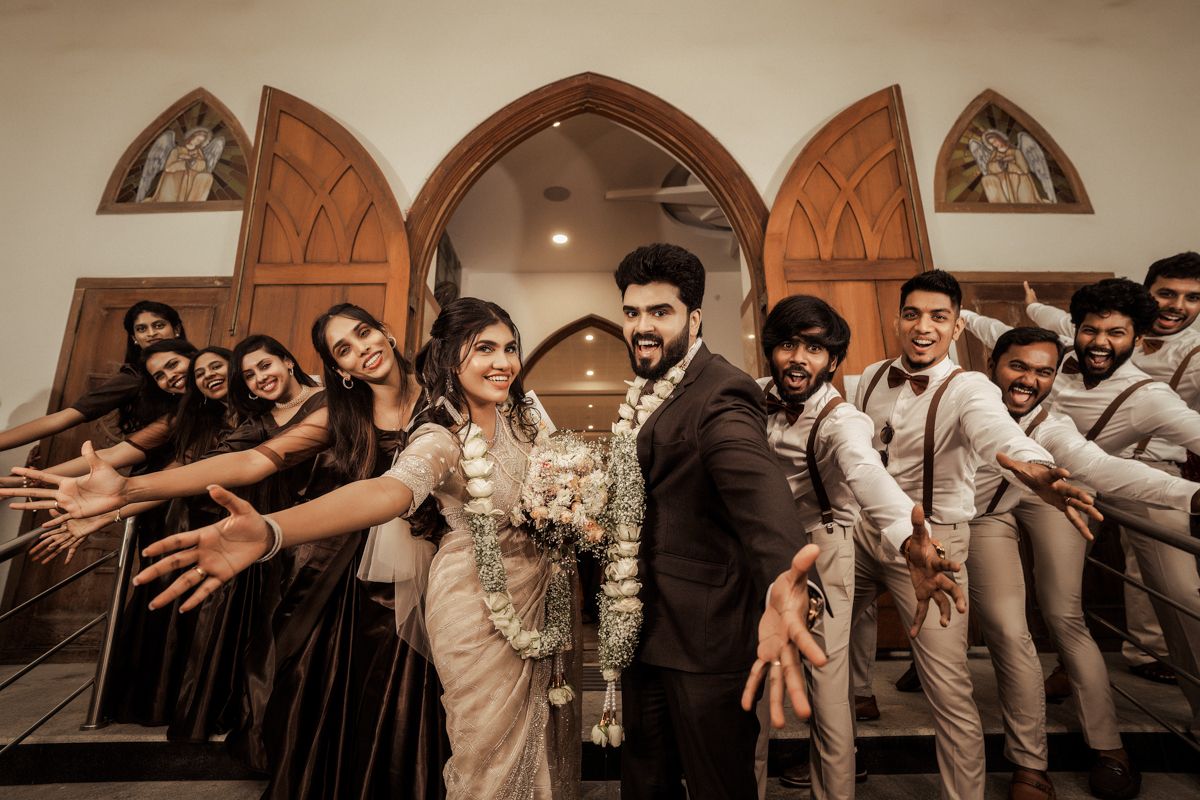 Wedding party in formal attire poses playfully in front of arched church entrance with warm lighting.