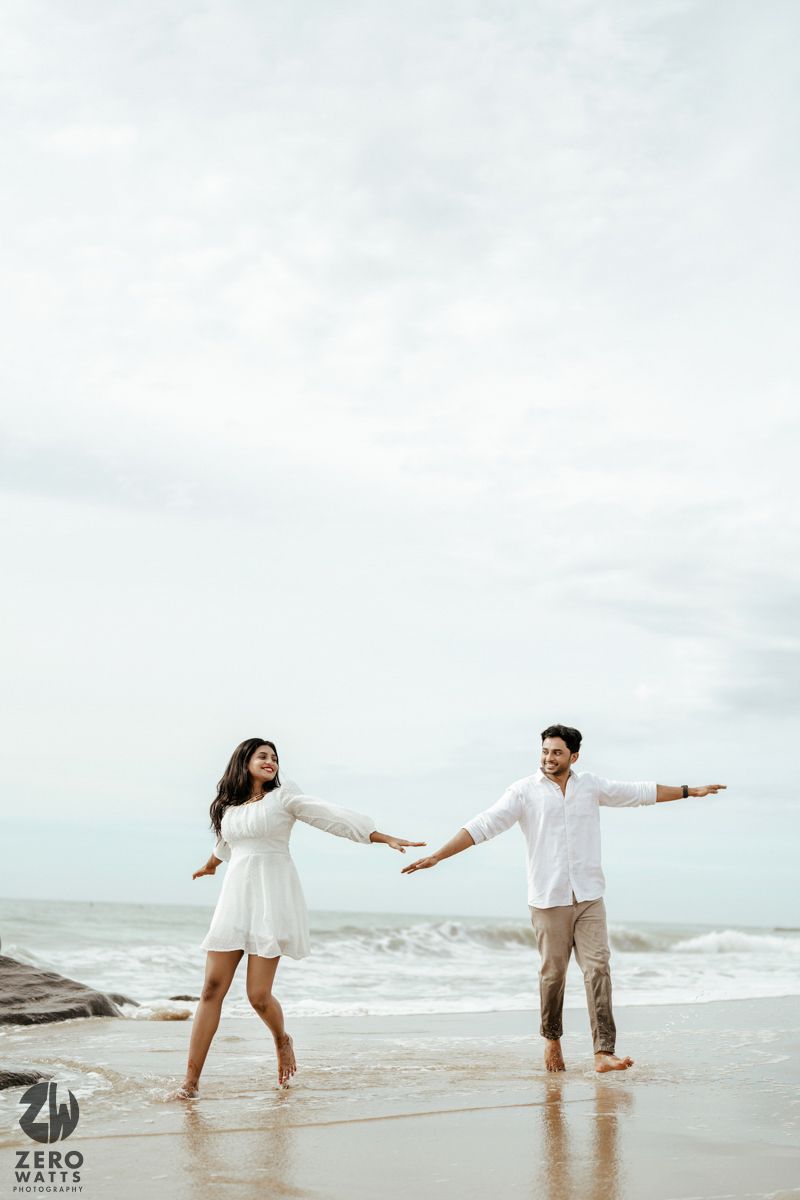 A couple in matching white outfits dances playfully at the edge of the ocean surf.