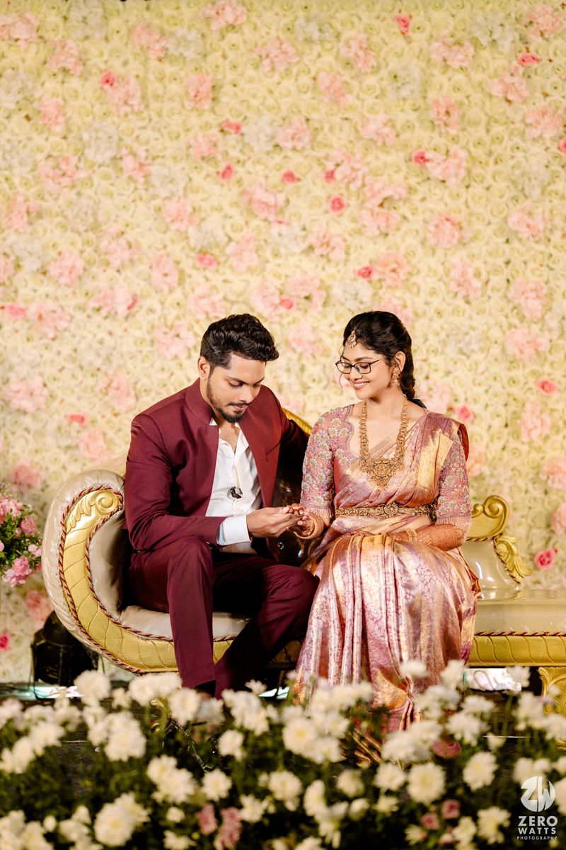 A couple sits together on an elegant gold couch against a floral backdrop in coordinating formal attire.