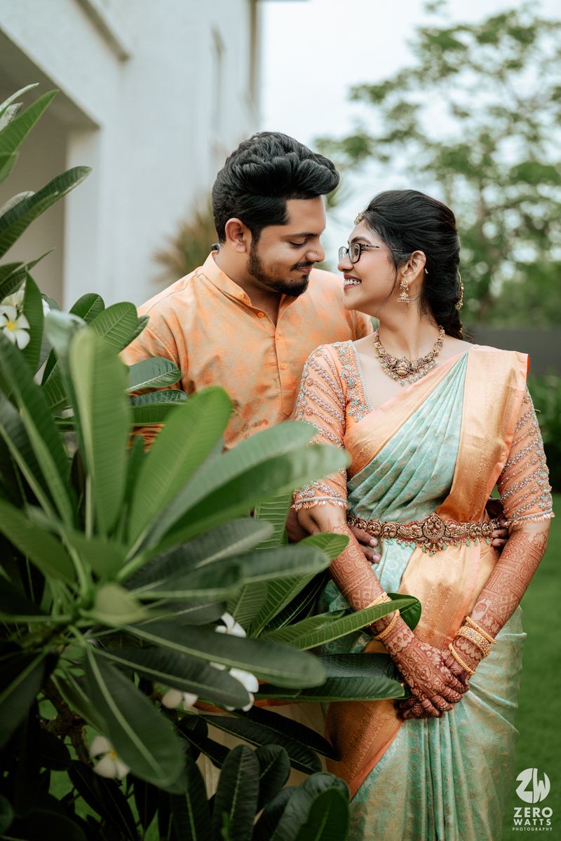 A couple in traditional South Indian attire pose intimately among tropical plants wearing peach and mint colored outfits.