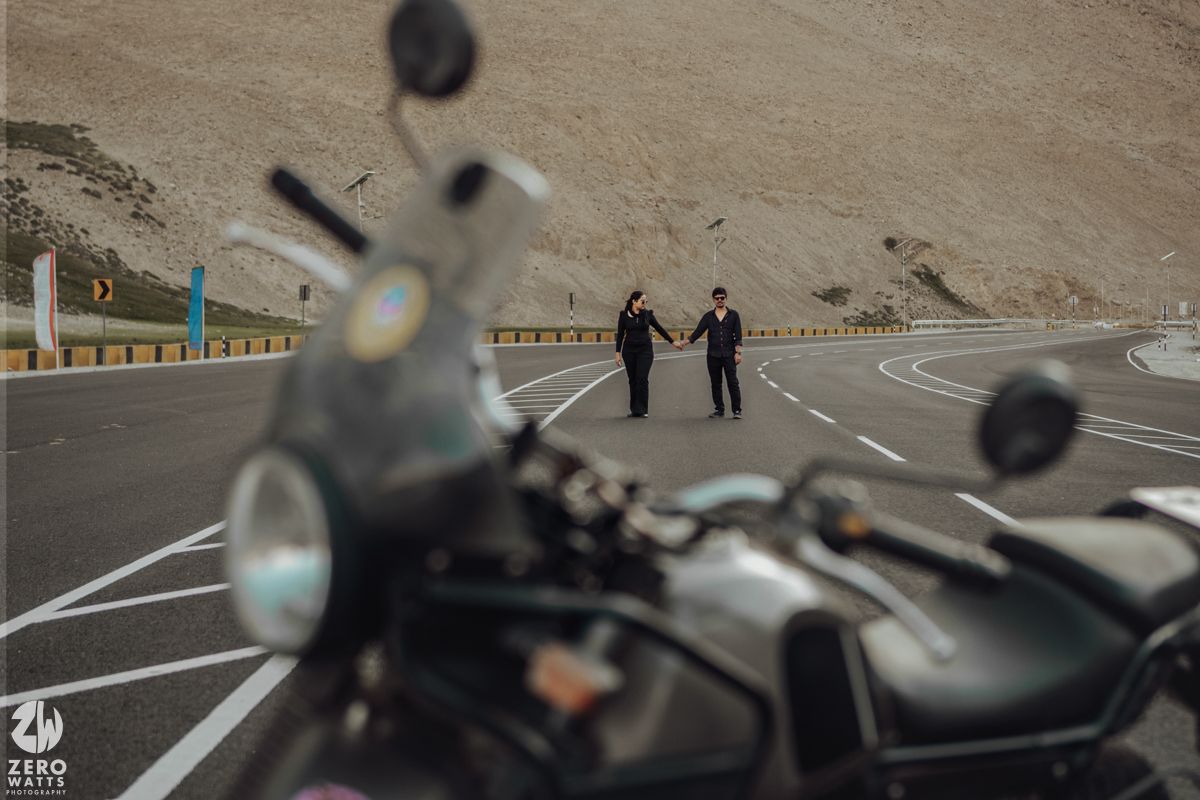 A motorcycle parked on a winding mountain road with two silhouetted figures walking in the distance.