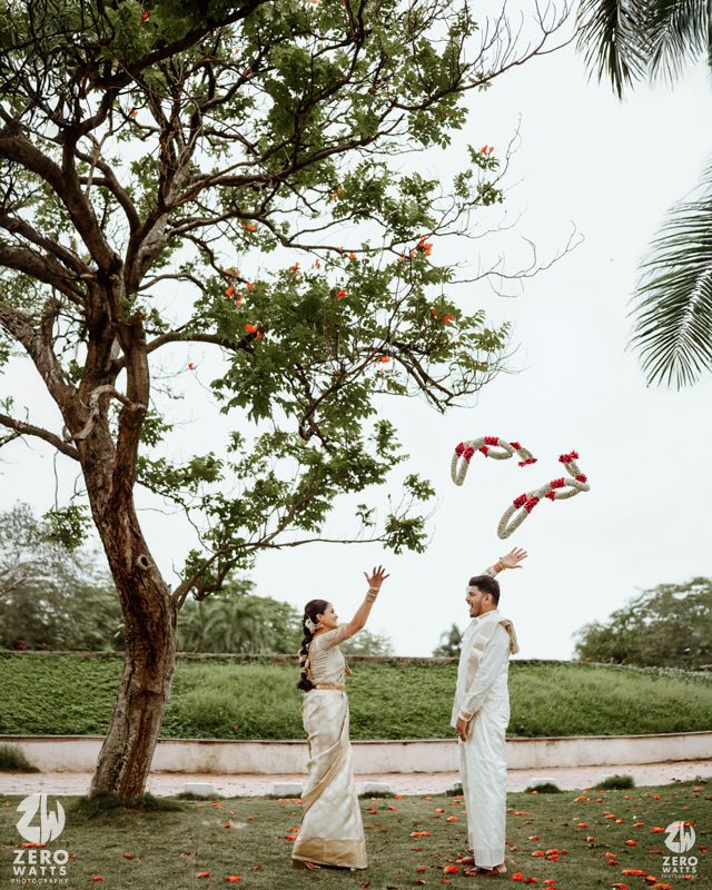 Two people in white attire throw red fabric in the air beneath a large tree in a garden setting.