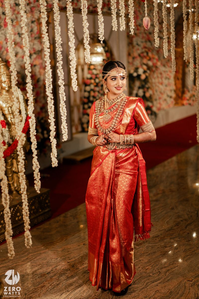 A bride in a striking red silk saree and traditional South Indian bridal jewelry stands beneath floral decor.