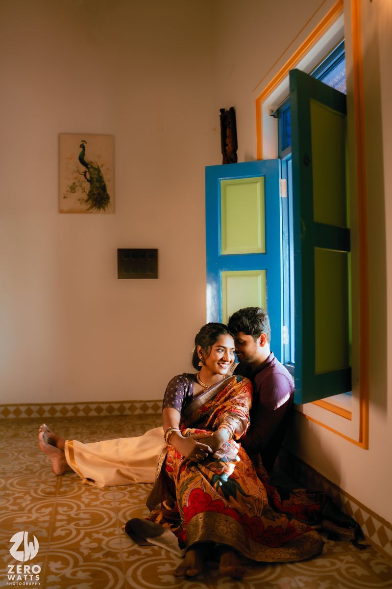 A couple sits intimately by a turquoise window frame in a room with patterned floors and peacock wall art.