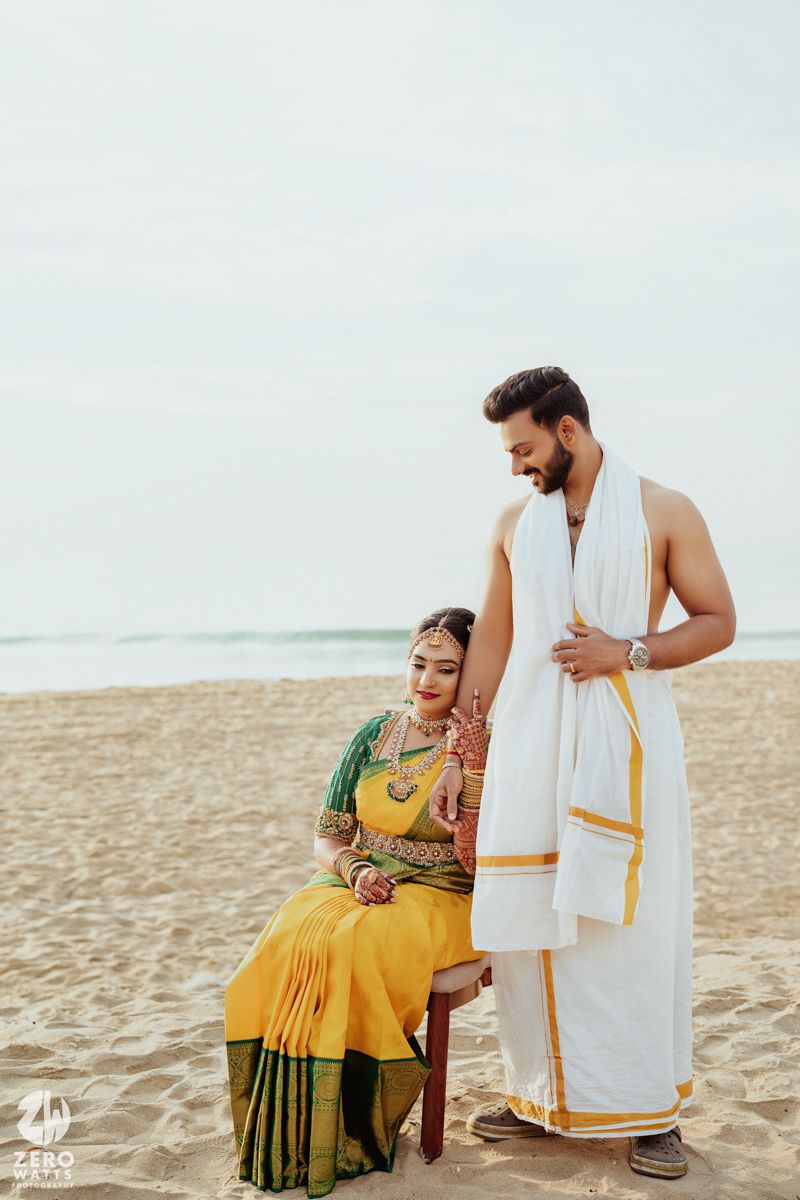 A couple in traditional Indian attire poses on a beach, with her in a yellow sari and him in a white dhoti.