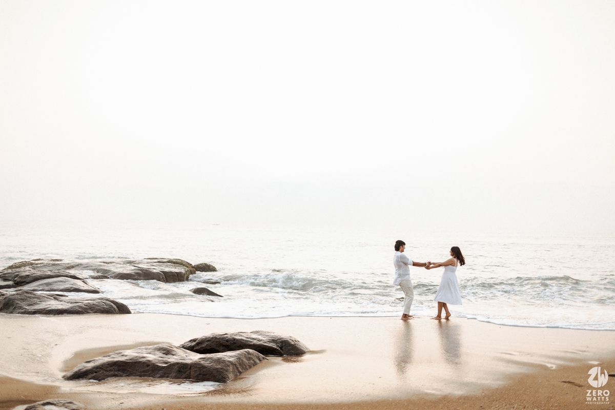 A couple dances together on a misty beach with rocky outcrops and gentle waves in soft evening light.