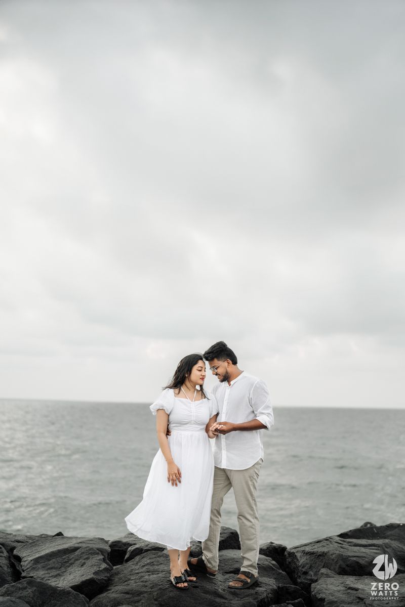 Romantic couple in white outfits standing on rocky beach under cloudy sky.