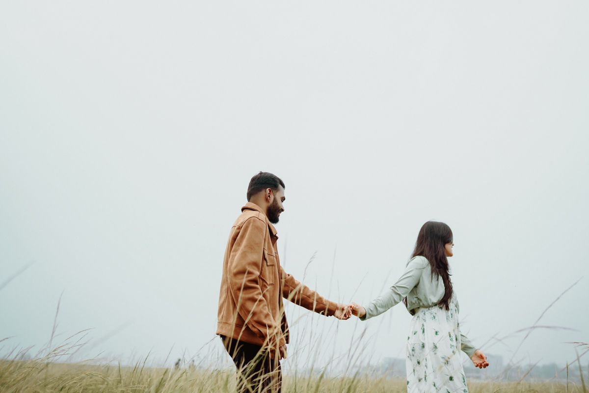Couple walking hand in hand through a misty field wearing brown jacket and white dress.