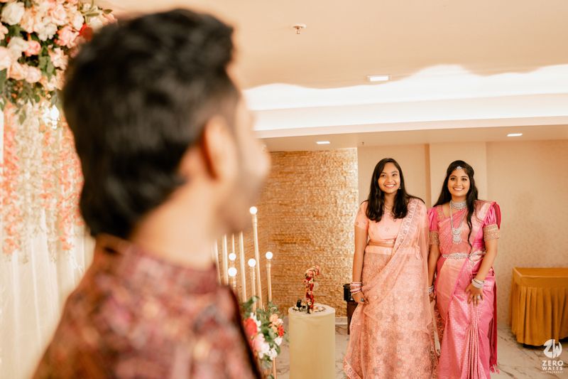 People in pink and coral traditional outfits attend an elegantly decorated indoor celebration with floral decor and candles.