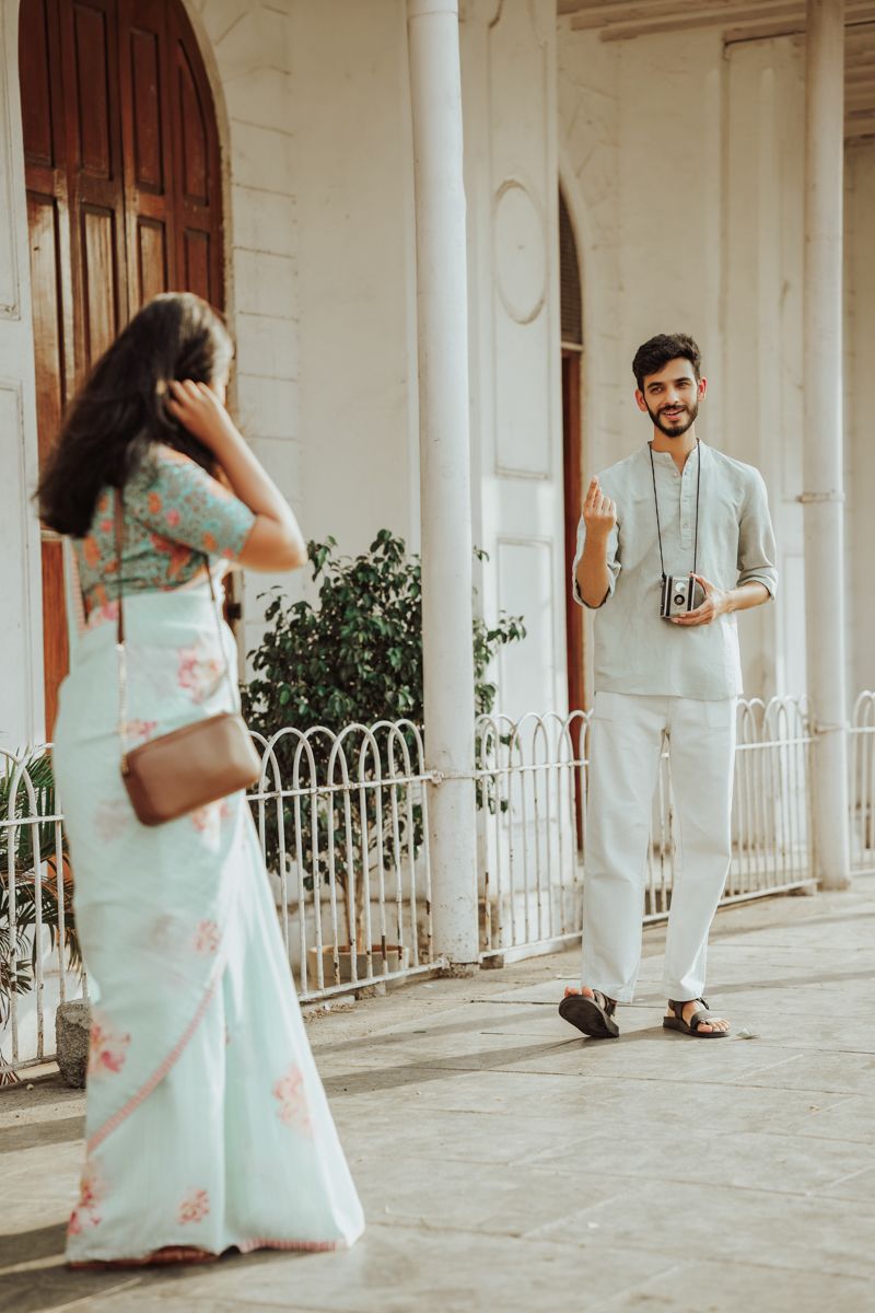 A couple in light colored outfits pose for photos in front of a colonial style building with pillars.