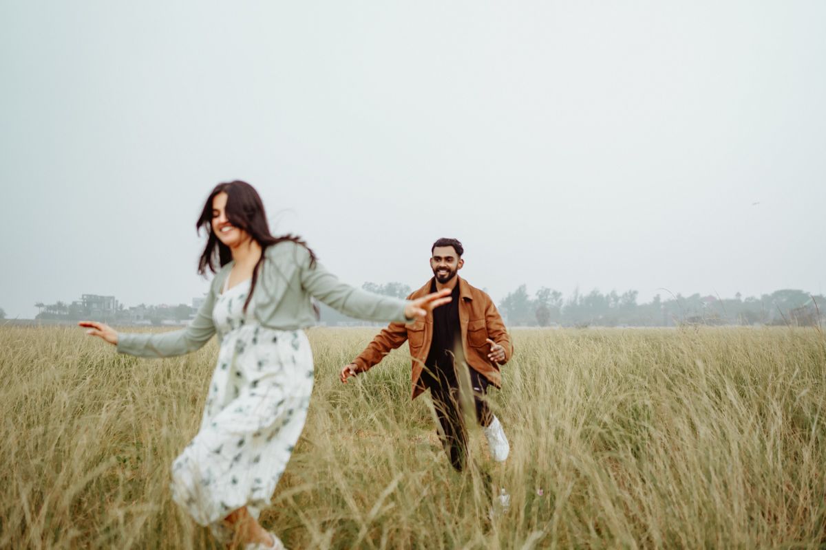 A couple runs joyfully through a tall grass meadow on an overcast day, with the person in front wearing a flowing dress.