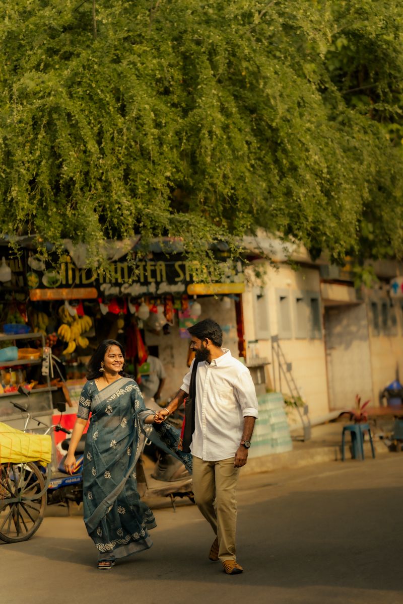 People walk past a street vendor under lush green trees in an urban Indian setting during golden hour.