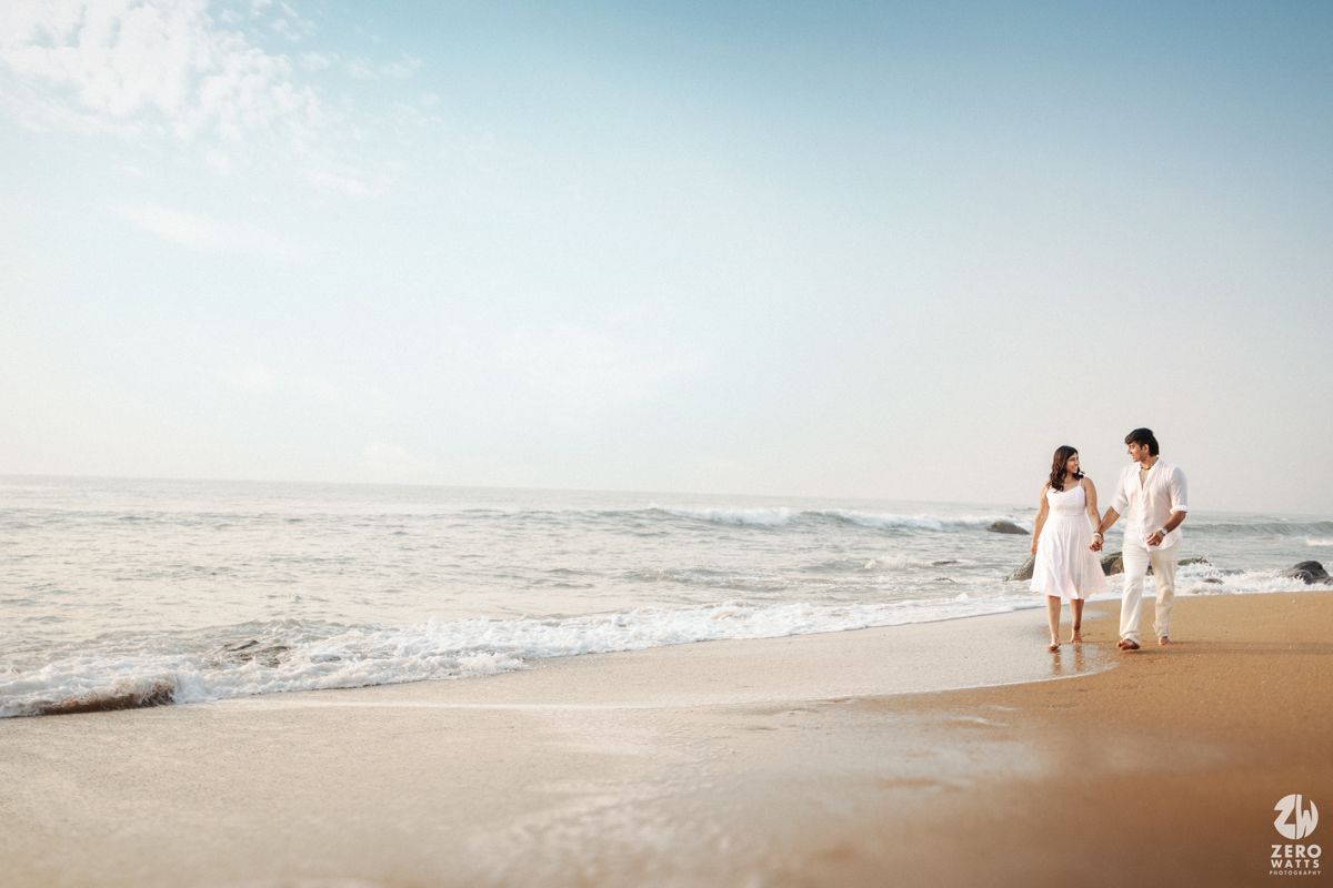 A couple in white attire takes a romantic stroll along a sandy beach shoreline.