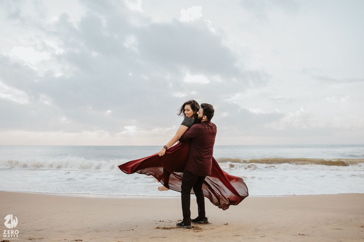 A romantic beach photoshoot with flowing maroon dress catching wind against cloudy sky.