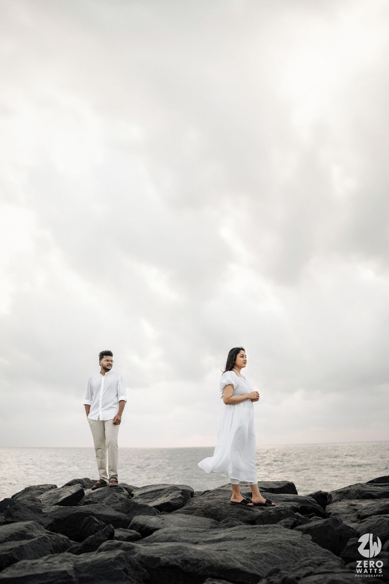 Couple in white outfits standing apart on black rocks by cloudy ocean.