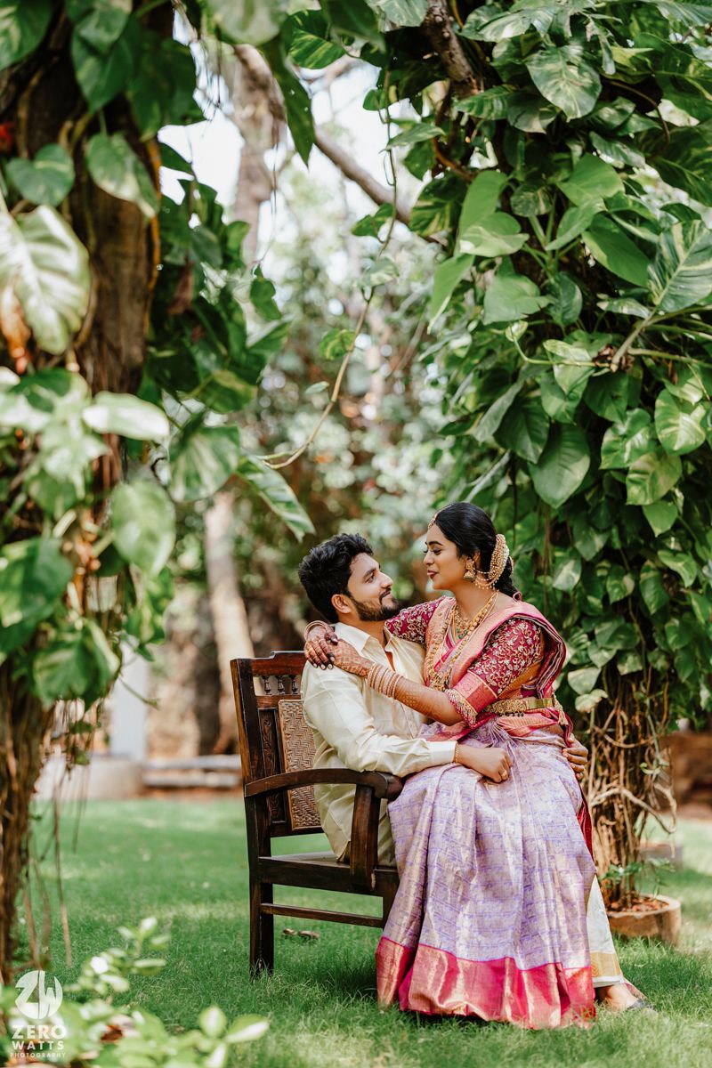 A couple shares an intimate moment on a wooden bench beneath lush green foliage in traditional Indian attire.