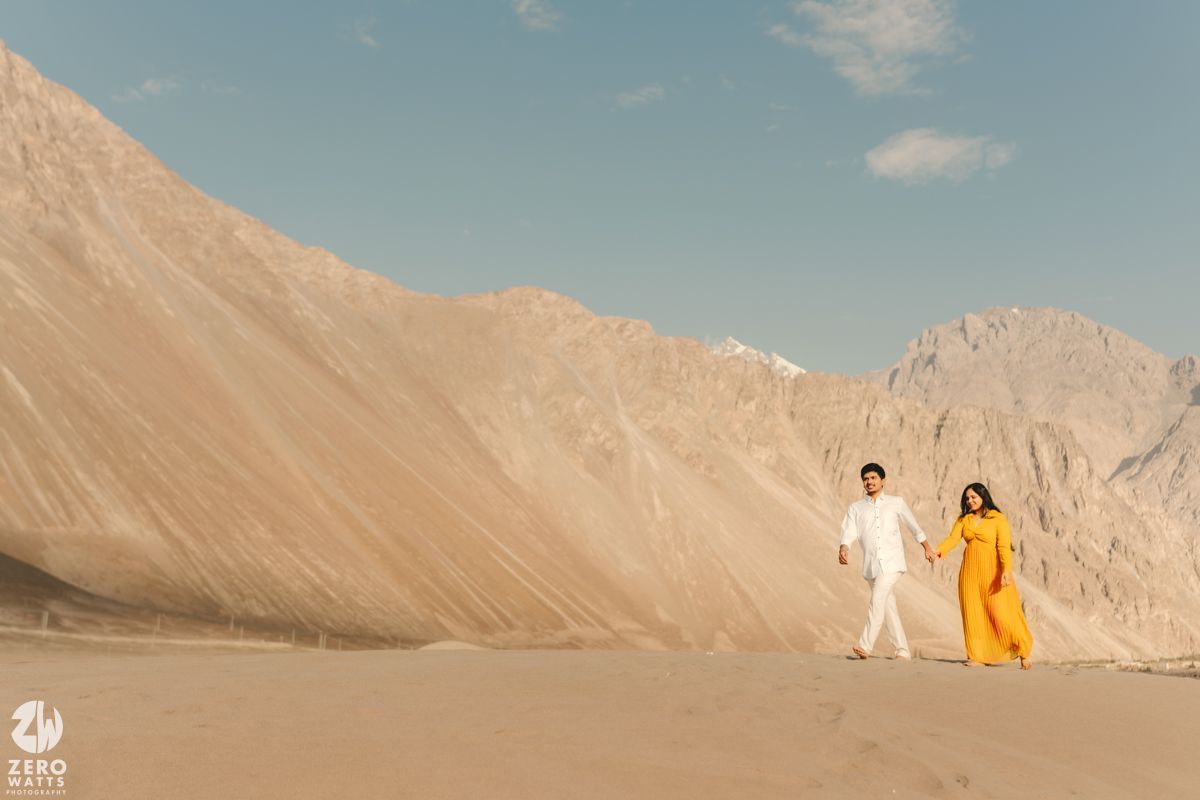 A couple in white and yellow outfits poses against a dramatic backdrop of desert mountains under a bright blue sky.