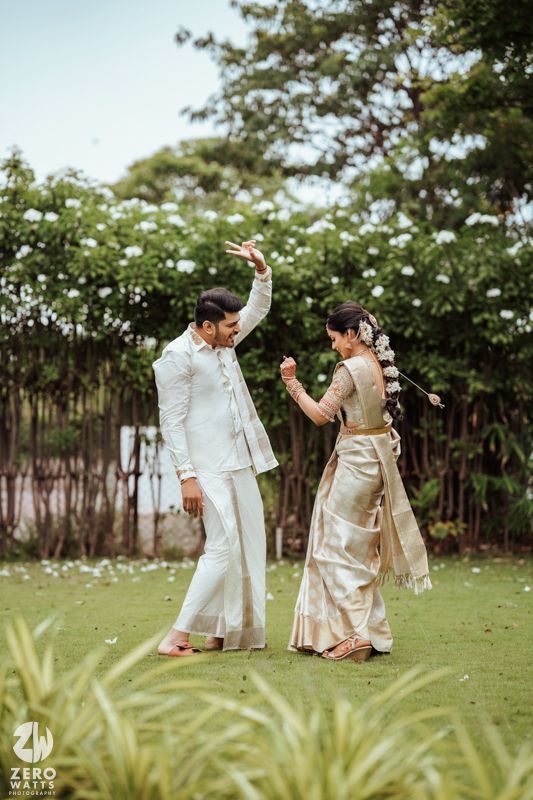 A couple in traditional Indian attire dancing joyfully on a lawn with greenery in the background.