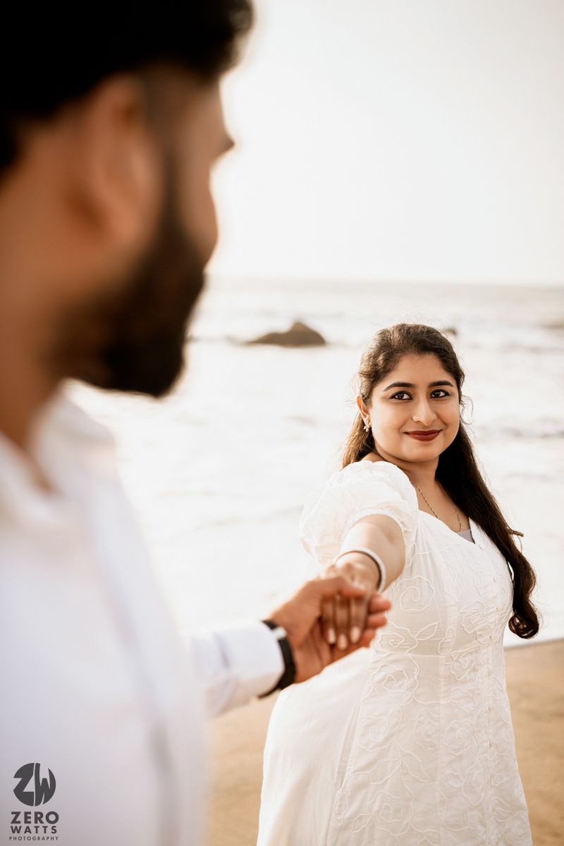 A romantic photoshoot on the beach with a woman in white dress holding hands with someone in the foreground.