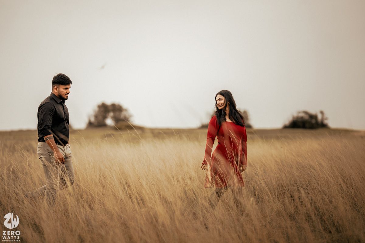 A couple stands apart in a wheat field wearing red dress and black shirt during sunset.