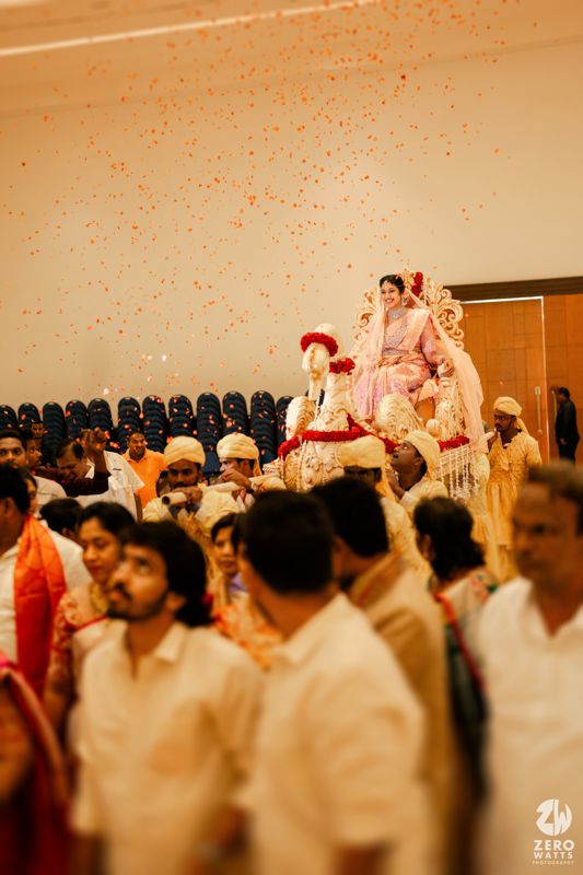 Wedding guests celebrate during a traditional Indian ceremony with vibrant rose petals showering down in an indoor venue.