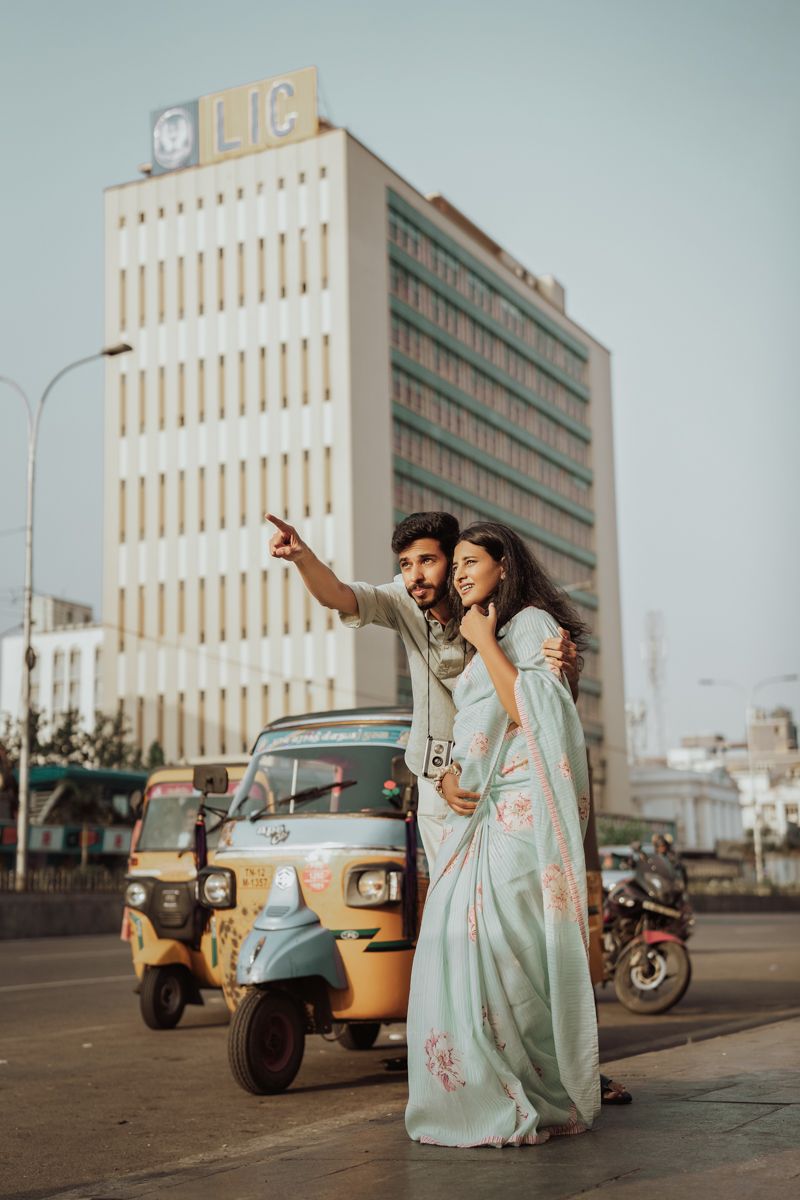 A couple in traditional Indian attire poses near an auto rickshaw in front of a tall city building.
