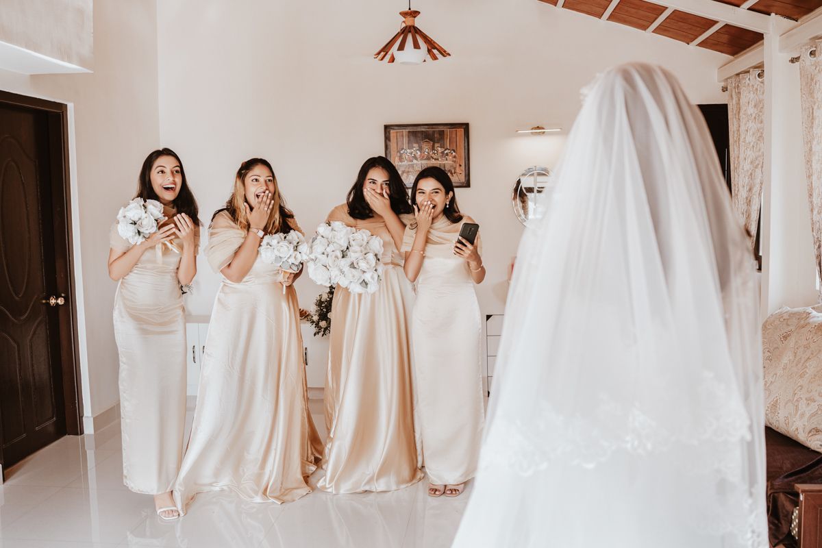 Wedding party in champagne bridesmaid dresses react with excitement to wedding gown hanging in bridal suite.