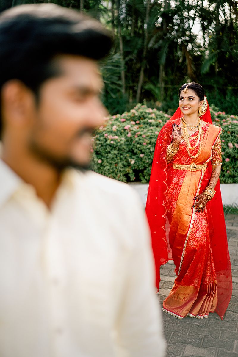 A traditional Indian wedding scene with a person in white clothing in foreground and someone in a vibrant red and gold sari.