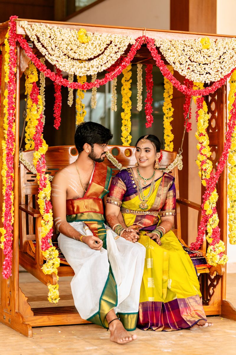 Couple seated under ornate mandap decorated with red and yellow flower garlands during traditional ceremony.