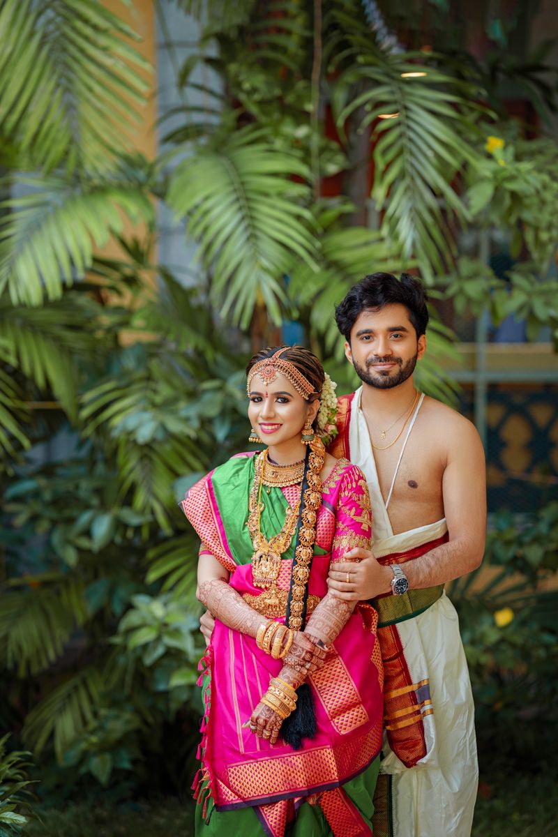 Newlywed couple in traditional South Indian wedding attire posing against lush tropical foliage backdrop.