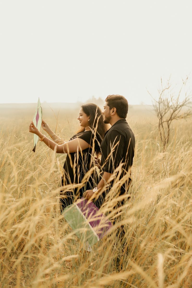 A couple stands together in a golden wheat field during sunset while taking a photo with a phone.