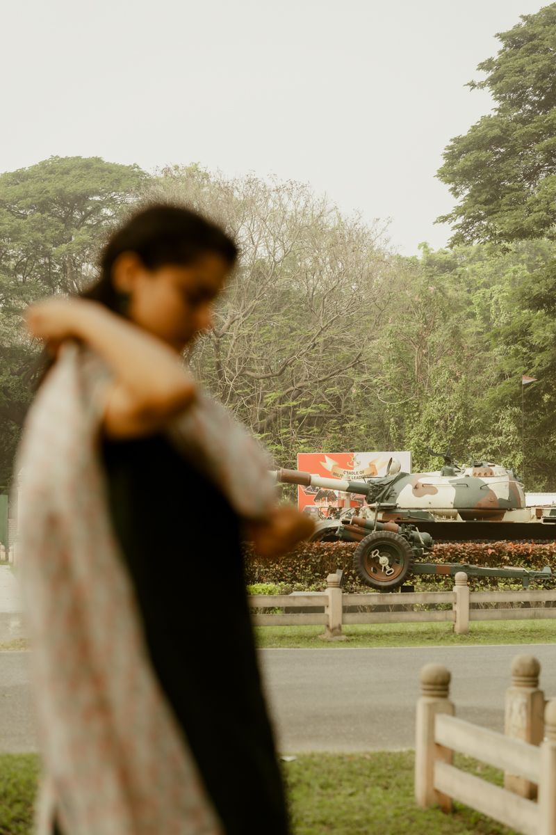 A figure in traditional dress stands in a garden setting with vehicles visible in the background.