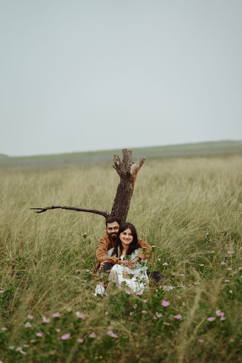A couple sits together in a grassy field near a bare tree trunk on a cloudy day.