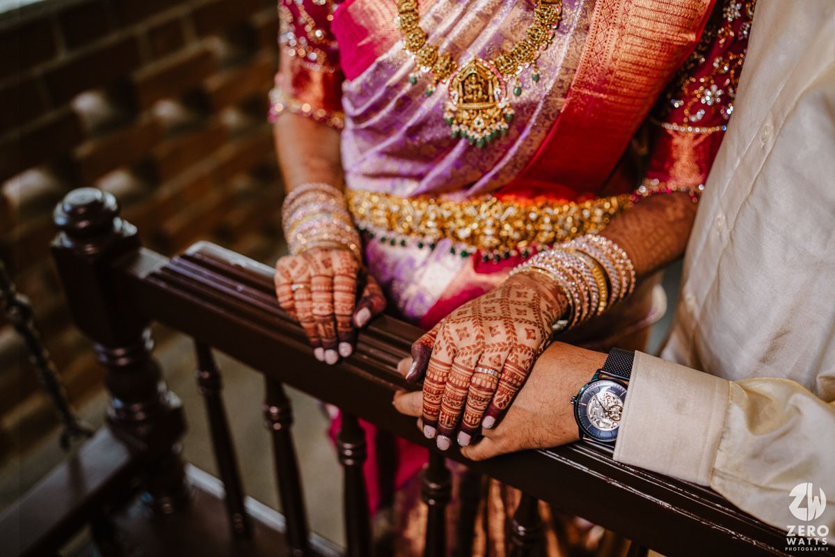 Detail shot of hennaed hands adorned with jewelry resting on wooden railing at an Indian wedding celebration.