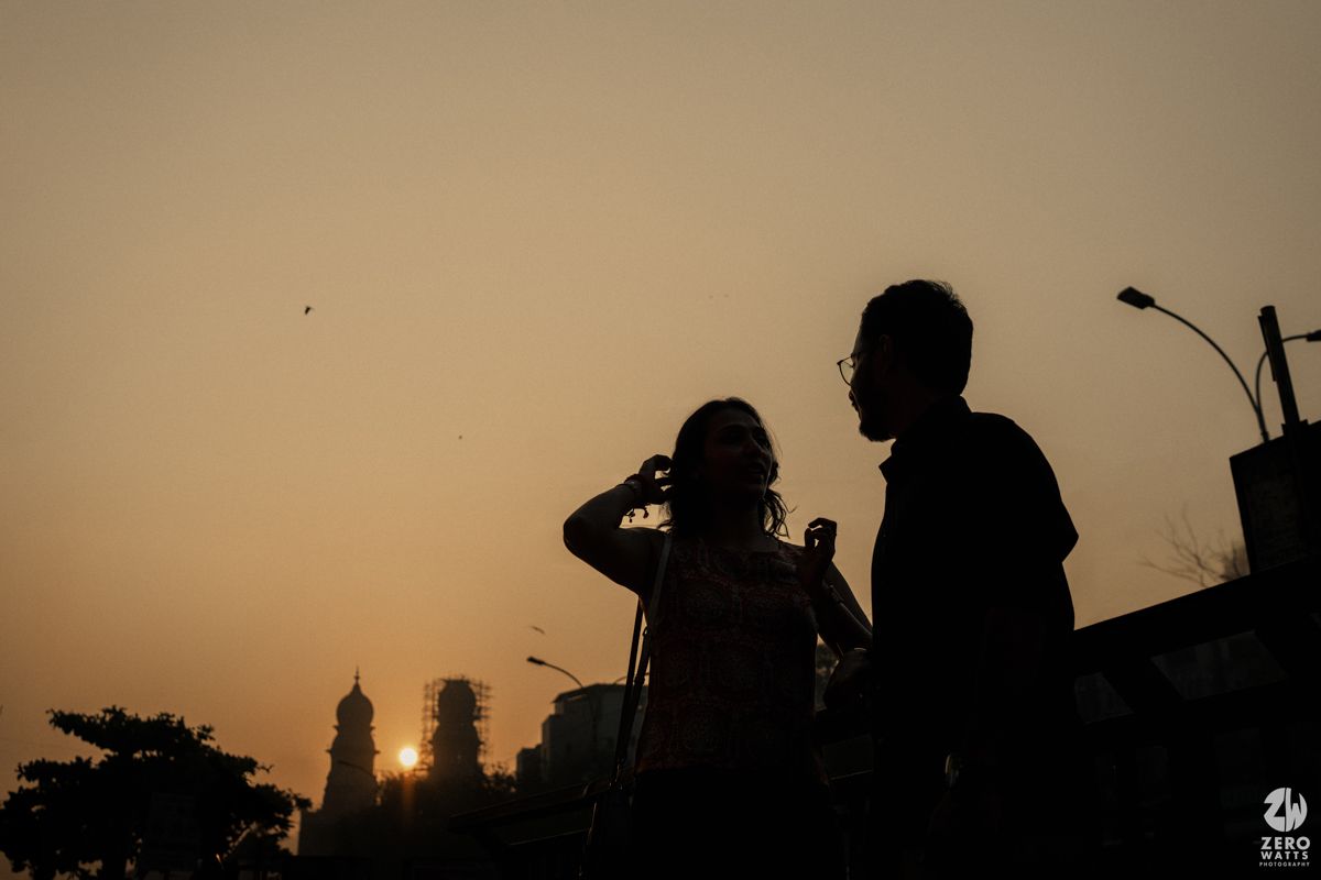 Two silhouettes stand against a warm sunset sky with city buildings visible in the background.