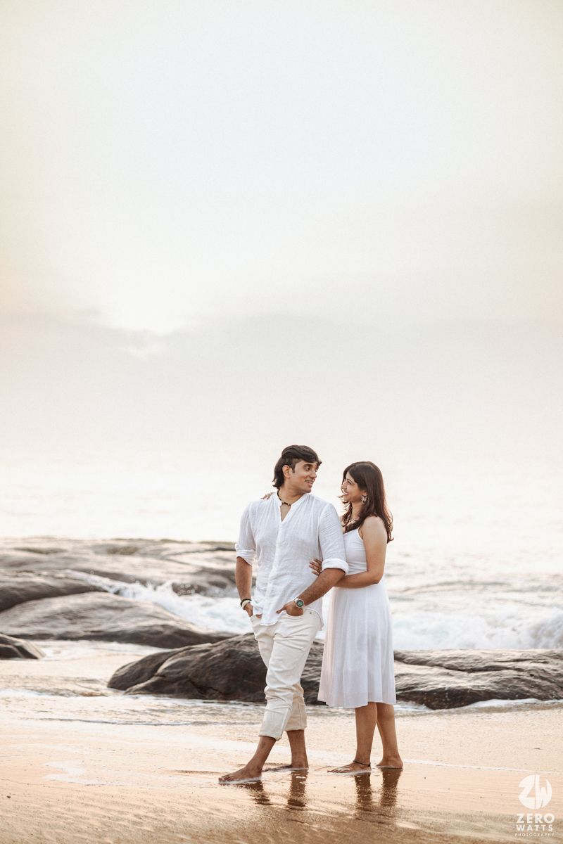 A couple in white outfits stands together on a beach at sunset near rocky formations by the ocean.