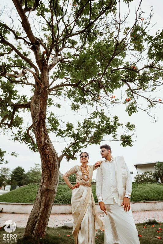 Candid wedding photography showing couple in traditional attire posing beneath large tree branches.