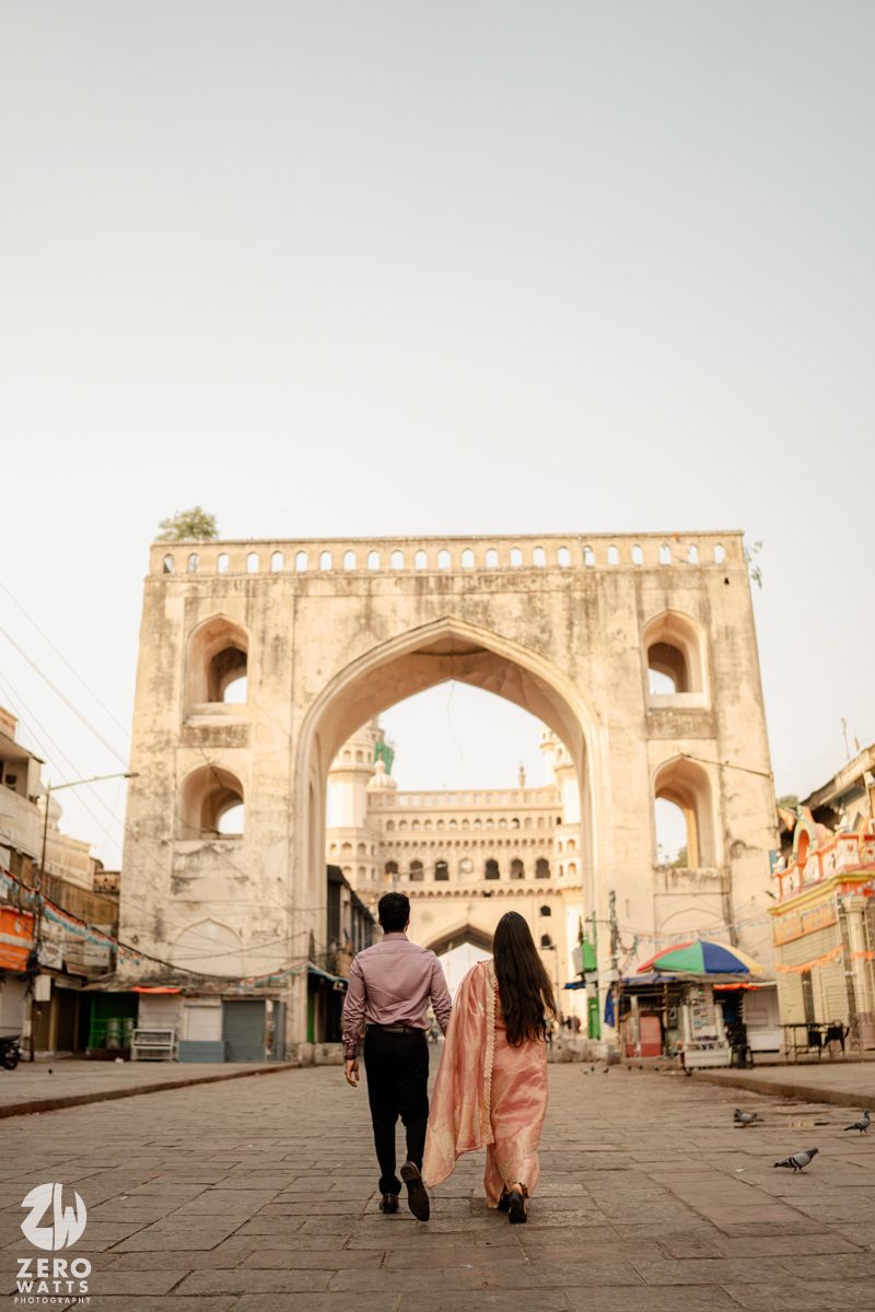 Romantic couple walks near historic sandstone archway architecture at sunset.