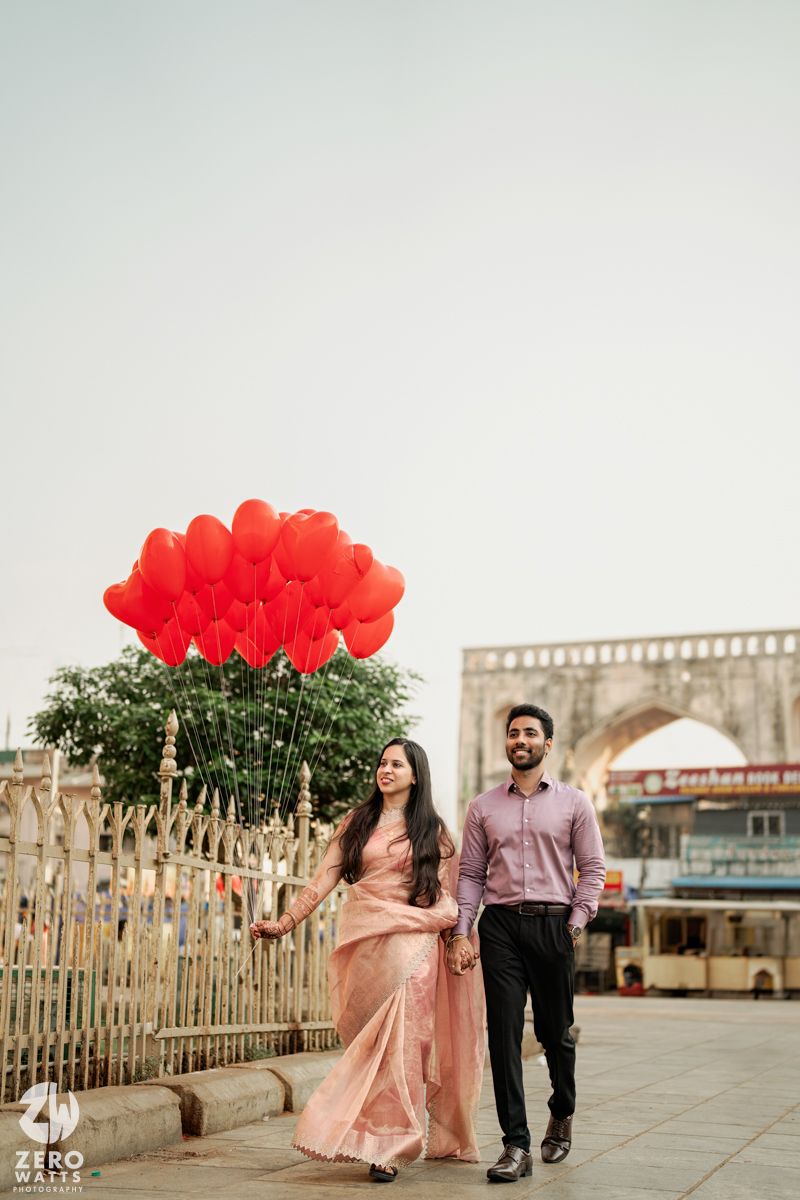 Romantic couple walking on a pier with red heart-shaped balloons against an urban backdrop.