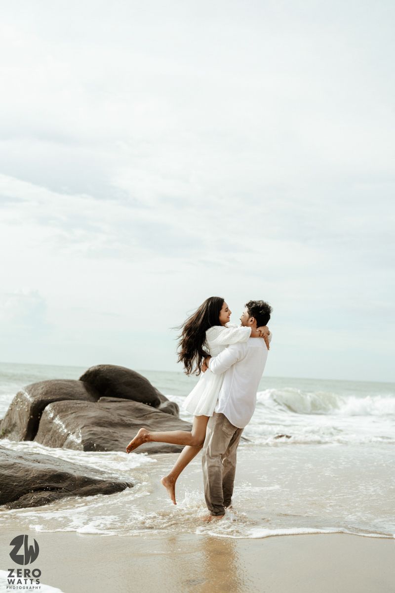 Playful couple embraces on rocky beach shore with waves crashing in the background.